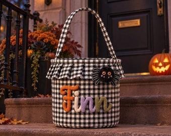 May include: A black and white gingham fabric bucket with a matching handle and a ruffled top edge. The front features the name Finn in colorful, textured, yarn-like letters and a small, fuzzy black spider with orange eyes attached to the side.