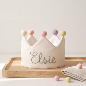 May include: A cream-colored fabric crown featuring the embroidered name Elsie in green cursive script. The crown is adorned with five small, round, felt pom-poms in pastel shades of white, peach, pink, lavender, and yellow along the top points.