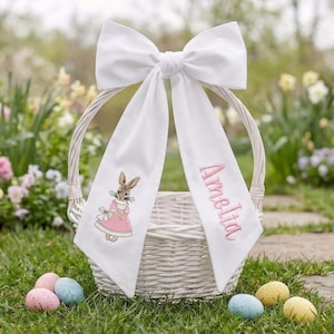 Op de afbeelding: Een witte rieten paasmand met een grote witte strik en een lang wit stoffen paneel. Het paneel is geborduurd met een konijn in een roze jurk en de naam "Amelia" in roze. Kleurrijke paaseieren liggen verspreid op het gras.