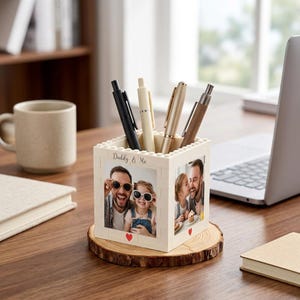 May include: A cream-colored pen holder with the text "Daddy & Me" and photos of a father and child. The holder contains several pens and sits on a wooden coaster. A coffee mug, notebook, and laptop are in the background.
