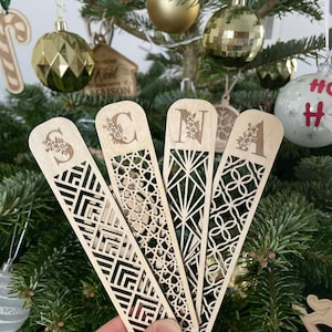 Peut inclure: Quatre marque-pages en bois forment le mot « SCAN » devant un sapin de Noël décoré. Chaque marque-page présente un motif géométrique différent et une lettre avec des motifs floraux. L'arbre est orné de boules dorées et miroitantes.