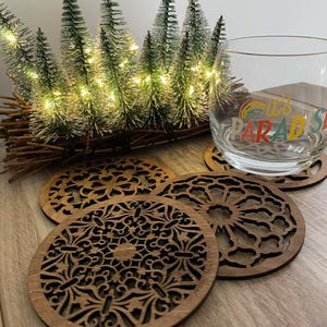 Könnte beinhalten: Vier aufwendig gestaltete Untersetzer aus Holz mit dunkler brauner Oberfläche werden gezeigt. Die Untersetzer weisen detaillierte, durchbrochene Muster auf. Ein klares Glas mit dem Text "it's PARADISE" steht auf einem der Untersetzer. Im Hintergrund befindet sich ein kleiner dekorativer Weihnachtsbaum.