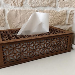 Peut inclure: Une boîte à mouchoirs rectangulaire en bois brun foncé avec un motif floral découpé au laser sur le dessus et les côtés. La boîte présente une ouverture centrale sur la surface supérieure par laquelle un mouchoir blanc est extrait.
