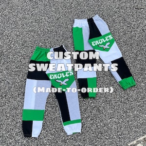 Può includere: Due paia di pantaloni della tuta personalizzati con un design patchwork. I pantaloni della tuta presentano pannelli verdi, neri, bianchi e grigi. Ogni gamba dei pantaloni ha una toppa verde con la scritta "EAGLES" e una grafica di un'aquila in volo. Viene visualizzato il testo "CUSTOM SWEATPANTS (MADE-TO-ORDER)".