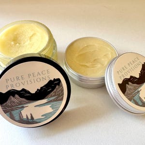 Könnte beinhalten: Draufsicht auf mehrere Balsambehälter. Einer befindet sich in einem transparenten Glas, zwei in kleinen Metalldosen. Die Deckel der Dosen zeigen ein Berg- und Seelandschaftsdesign mit dem Text "PURE PEACE PROVISIONS."