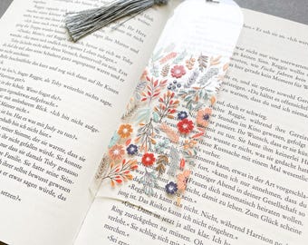 May include: A clear bookmark with a floral design and a grey tassel sits on an open book. The bookmark displays colourful flowers and leaves in red, orange, and blue. The book's pages are visible.