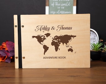May include: A wooden adventure book with the names "Ashley & Thomas" and a world map design in brown. The words "ADVENTURE BOOK" are printed below the map. The book has a black spine and is on a wooden surface.