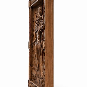 May include: A detailed, wooden relief carving depicting a classical female figure in a framed rectangular panel. The relief is carved in a warm brown tone, with intricate details in the figure's hair, clothing, and the surrounding floral elements.