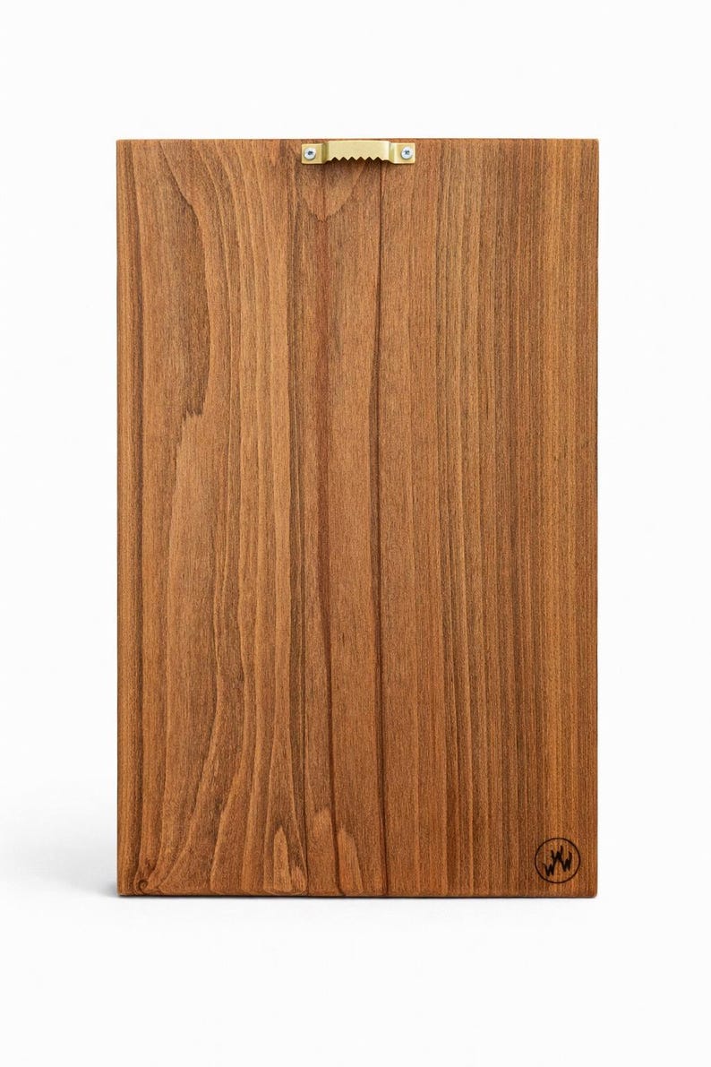 May include: A rectangular wooden board with a warm brown finish and vertical wood grain. A brass sawtooth hanger is attached to the top. A small circular logo is in the bottom right corner.