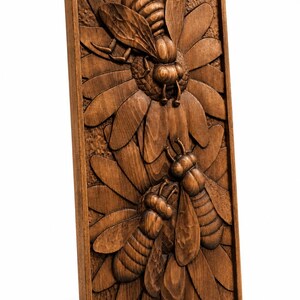 May include: A wooden relief carving of two bees on a flower. The bees are finely detailed, with visible wings and body segments. The artwork is framed in a rectangular shape and has a natural wood finish.