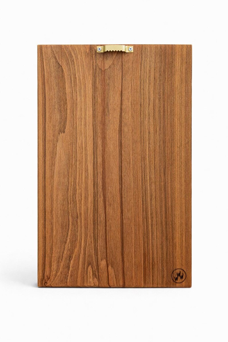 May include: A rectangular wooden board with a warm brown finish and vertical wood grain. A brass sawtooth hanger is attached to the top. A small circular logo is in the bottom right corner.