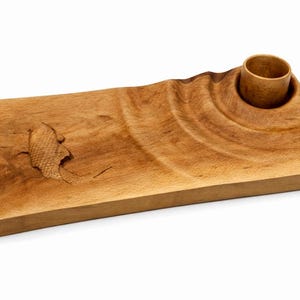 Puede incluir: Tabla de servir de madera hecha a mano con acabado natural. Presenta un diseño tallado y una hendidura circular para una taza pequeña. La veta de la madera es visible. La tabla tiene forma rectangular.