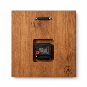 May include: Back of a square wooden clock with a visible clock mechanism. The wood has a warm brown tone. A black metal hanger is attached at the top, and the clock mechanism is visible in a recessed square. A logo is in the bottom right corner.