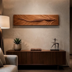 May include: A detailed wooden carving of an alligator mounted on a wall. The sculpture features textured scales and a long body. It is displayed above a dark wood cabinet, with a plant and a small statue on top.