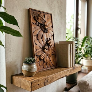 May include: A carved wooden wall plaque depicting bees on a flower, displayed on a reclaimed wooden shelf. The shelf also holds a small potted succulent and books. The artwork and shelf are in natural wood tones, creating a rustic aesthetic. The shelf is approximately 30 cm deep.