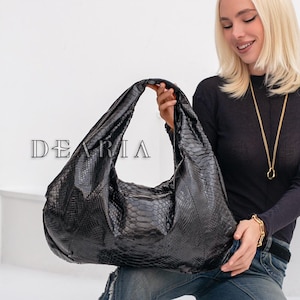 May include: A large black handbag with a textured, crocodile-like pattern. The bag is held by a person wearing a black long-sleeve shirt and blue jeans. The bag has the text "DEARIA" on it.