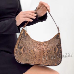 Könnte beinhalten: Braune und beige Schultertasche mit Schlangenmuster und geschwungener Form. Die Tasche hat einen passenden braunen Riemen. Der Text "PYTOSHASHOP" ist auf der Tasche zu sehen. Die Tasche wird von einer Person gehalten.