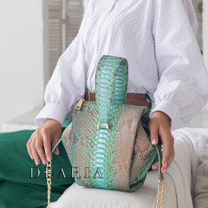 May include: A teal green and brown snakeskin handbag with a gold chain strap. The bag has a unique, geometric shape and is sitting on a white sofa.