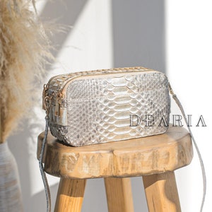 Könnte beinhalten: Eine rechteckige, hellfarbene Crossbody-Tasche aus Schlangenleder mit goldenem Reißverschluss und Kettenriemen liegt auf einem Holzschemel. Auf der Tasche steht das Wort "DEARIA". Im Hintergrund eine Vase mit getrockneten Pflanzen.