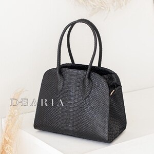 Könnte beinhalten: Schwarze Handtasche mit strukturierter Schlangenmusterung. Die Tasche hat zwei runde Henkel und eine strukturierte Form. Das Wort "DEARIA" ist auf der Seite aufgedruckt. Die Tasche steht auf einer weißen Oberfläche. Länge ca. 25 cm.