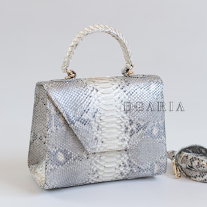 May include: A silver and white snakeskin handbag with a top handle and a detachable shoulder strap. The bag has a flap closure and gold-coloured hardware. The word "DEARIA" is printed on the front of the bag.