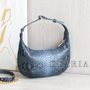 May include: A blue snakeskin-effect handbag with a curved design and a gold zip. The bag has a matching strap and the word "DEARIA" is printed on the side. The bag is on a white surface.