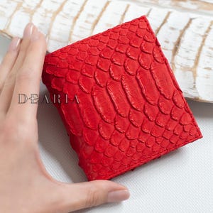 Può includere: Un portafoglio rosso vivo con un motivo strutturato a forma di scaglie. Il portafoglio è quadrato e sembra essere in pelle. La parola "DEARIA" è stampata sul portafoglio. Il portafoglio è tenuto da una mano.