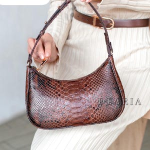 Könnte beinhalten: Eine braune Handtasche mit Schlangenmuster, geschwungener Form und Reißverschluss. Die Tasche hat einen braunen Riemen und goldfarbene Beschläge. Der Text "size S" befindet sich in einem weißen Feld oben links. Das Wort "DEARIA" ist auf der Tasche aufgedruckt.