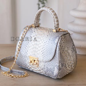 Peut inclure: Un sac à main argenté avec un motif en peau de serpent texturé. Il possède une poignée supérieure et une bandoulière en chaîne dorée. Le rabat avant est sécurisé par un fermoir doré. Le mot "DEARIA" est imprimé sur le devant du sac.