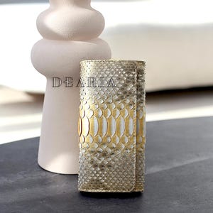 May include: A rectangular key case featuring a metallic gold and silver snakeskin pattern with a textured finish. The word DEARIA is printed in black capital letters on the front surface of the item. The key case has a vertical orientation.