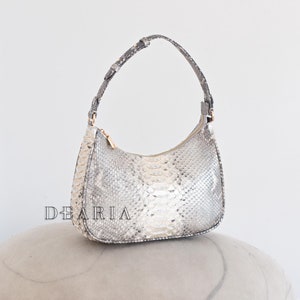 May include: A small, gray and white snakeskin handbag with a short handle. The bag is sitting on a gray, textured surface.