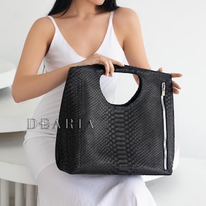 May include: Black handbag with a unique handle design and a zipper closure. The bag has a snakeskin pattern and the word "DEARIA" is printed on the front. The bag is held by a person wearing a white dress.