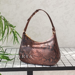 May include: A brown, snake-skin patterned handbag with a curved shape and a single shoulder strap. The bag has a gold zipper and hardware. The word "DBARIA" is visible on the bag. The bag is displayed on a white tiled surface.