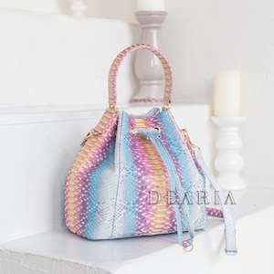 May include: A multi-colored bucket bag with a braided handle and a detachable shoulder strap. The bag features a pastel rainbow snakeskin pattern with the text "DEARIA" on the side. The bag is sitting on a white surface.