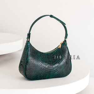 May include: A dark green, crescent-shaped handbag with a textured snakeskin pattern. The bag has a gold zip and a matching strap. The brand name "DE ARIA" is printed on the side.