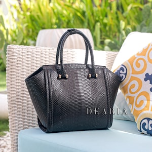 May include: Black leather handbag with a textured snakeskin pattern, featuring two top handles and gold-toned hardware. The bag is sitting on a light blue cushion. The word "DEARIA" is printed on the bag.