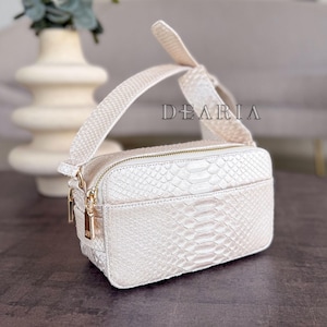 Könnte beinhalten: Cremefarbene rechteckige Umhängetasche mit Schlangenmuster. Die Tasche hat einen goldenen Reißverschluss und Beschläge sowie einen breiten, passenden Riemen. Das Wort "DEARIA" ist auf dem Riemen aufgedruckt.