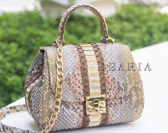 May include: A snakeskin handbag with a gold chain strap and handle. The bag features a gold clasp and a central stripe of lighter-coloured material. The word "DEARIA" is printed on the front of the bag.