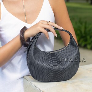 May include: A black, crescent-shaped handbag with a textured snakeskin pattern. The bag has a curved top handle and a gold zip. The word "DEARIA" is printed on the side of the bag.