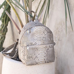 May include: A gray snakeskin-patterned backpack with silver zippers and the word "DEARIA" printed on the front. The backpack has a top handle and adjustable straps. It is displayed outdoors near a potted plant.