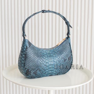 May include: A blue snakeskin-patterned handbag with a curved design and a top handle. The bag has a gold zip and the word "DEARIA" is printed on the side. It is displayed on a white table.