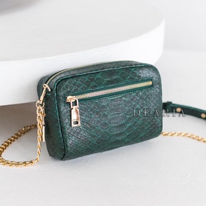 Könnte beinhalten: Dunkelgrüne Umhängetasche mit Schlangenmuster. Sie hat einen goldfarbenen Kettenriemen und einen Reißverschluss. Der Markenname "DEARIA" ist sichtbar. Die Tasche ist rechteckig.
