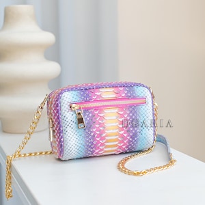 Könnte beinhalten: Eine rechteckige, mehrfarbige Umhängetasche mit einem goldenen Kettenriemen. Die Tasche hat einen Reißverschluss und ein lebendiges Muster aus Rosa, Lila, Blau und Gelb. Der Text "DEARIA" ist auf der Seite der Tasche zu sehen.