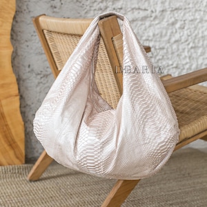 May include: A light beige snakeskin tote bag with a slouchy design. The bag is hanging on a wooden chair with a woven seat.