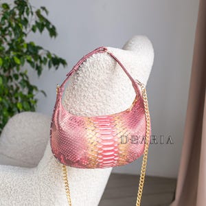 May include: A pink and gold snakeskin-patterned handbag with a gold chain strap. The bag is crescent-shaped and features the brand name "DEARIA" in white text. The bag is resting on a white, textured chair.
