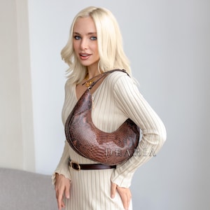 May include: A brown, crescent-shaped handbag with a snakeskin pattern. The bag is worn over the shoulder. The word "DEARIA" is printed on the bag. The woman is wearing a cream-colored dress and a brown belt.