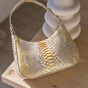 Könnte beinhalten: Eine hellgelbe und weiße Handtasche mit geschwungener Form und kurzem Riemen. Die Tasche hat ein Schlangenmuster und das Wort "DEARIA" ist auf der Seite aufgedruckt. Die Tasche steht auf einer Holzoberfläche.