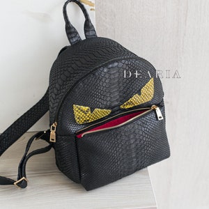 May include: Black backpack with a reptile skin texture, featuring yellow and black eye-shaped accents. The bag has a zippered front pocket with a gold zip and a red interior. The brand name "DEARIA" is printed on the bag.