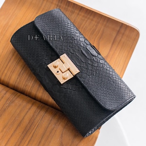 Könnte beinhalten: Schwarze Clutch mit Schlangenlederstruktur und goldfarbenem Verschluss. Das Wort "DEARIA" ist auf der Vorderseite aufgedruckt. Die Clutch liegt auf einem Holzstuhl.