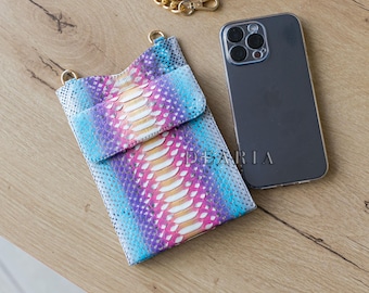 Puede incluir: Una funda de teléfono con estampado de piel de serpiente multicolor con una correa a juego y una cadena dorada. La bolsa es en tonos de azul, morado y rosa, con la palabra "DEARIA" impresa en la parte delantera. También se ve una funda transparente para el teléfono.